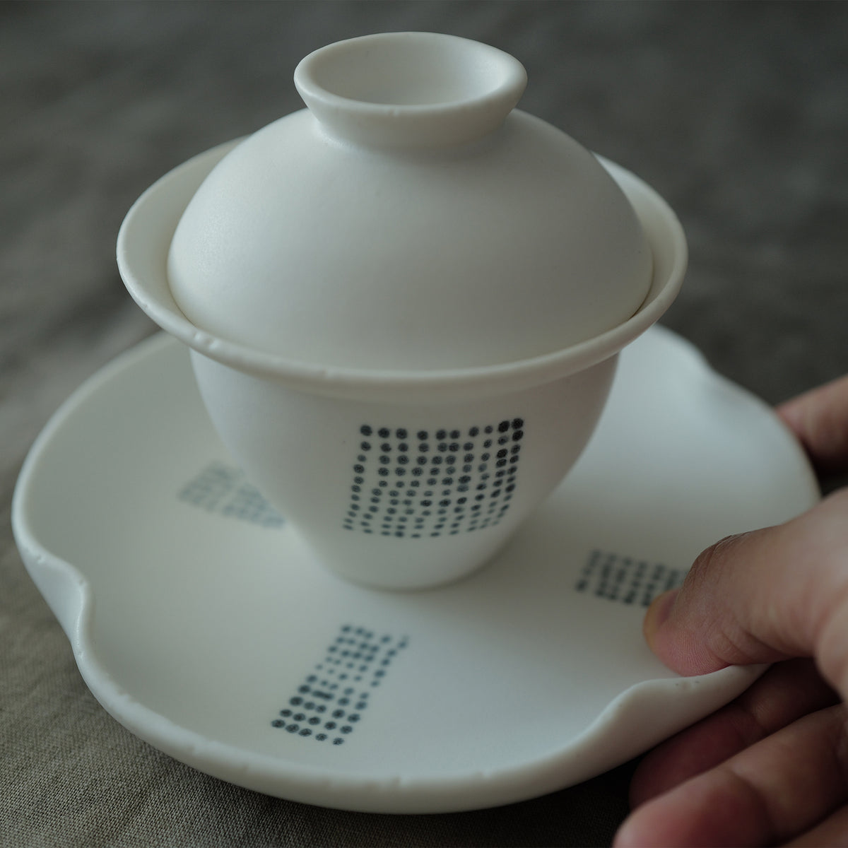 White ceramic gaiwan with lid on a matching saucer held by a hand.