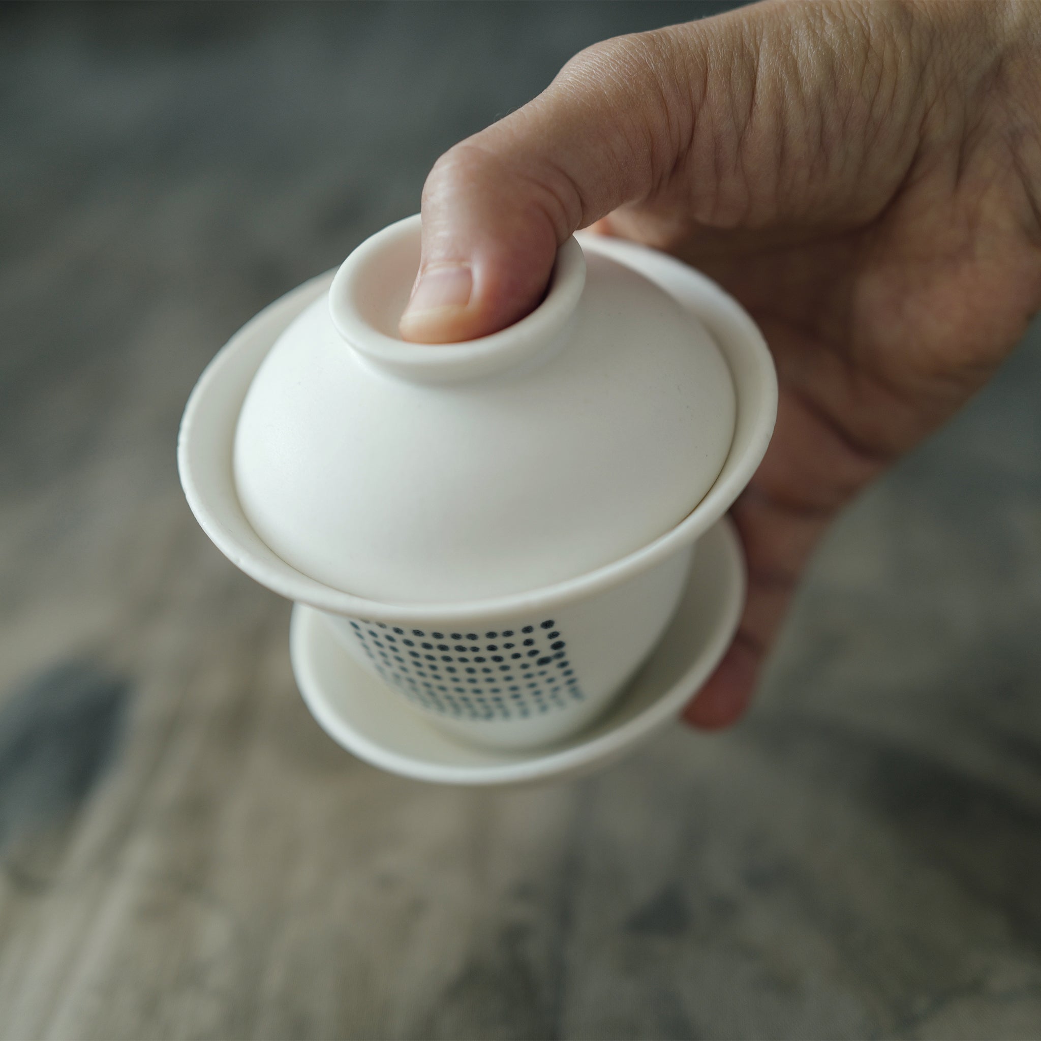 Hand holding a white ceramic gaiwan with a lid on a blurred background.