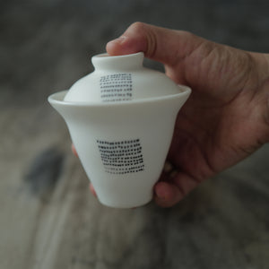 Hand holding a white ceramic gaiwan with dotted design against a blurred background.