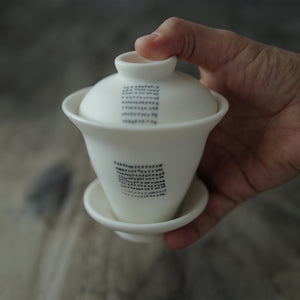 White ceramic gaiwan held by a hand against a blurred background.