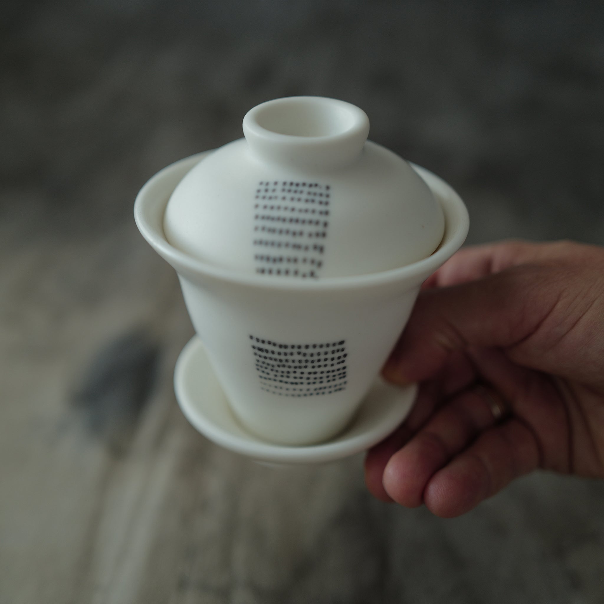 White ceramic gaiwan with a lid held by a hand against a blurred background.