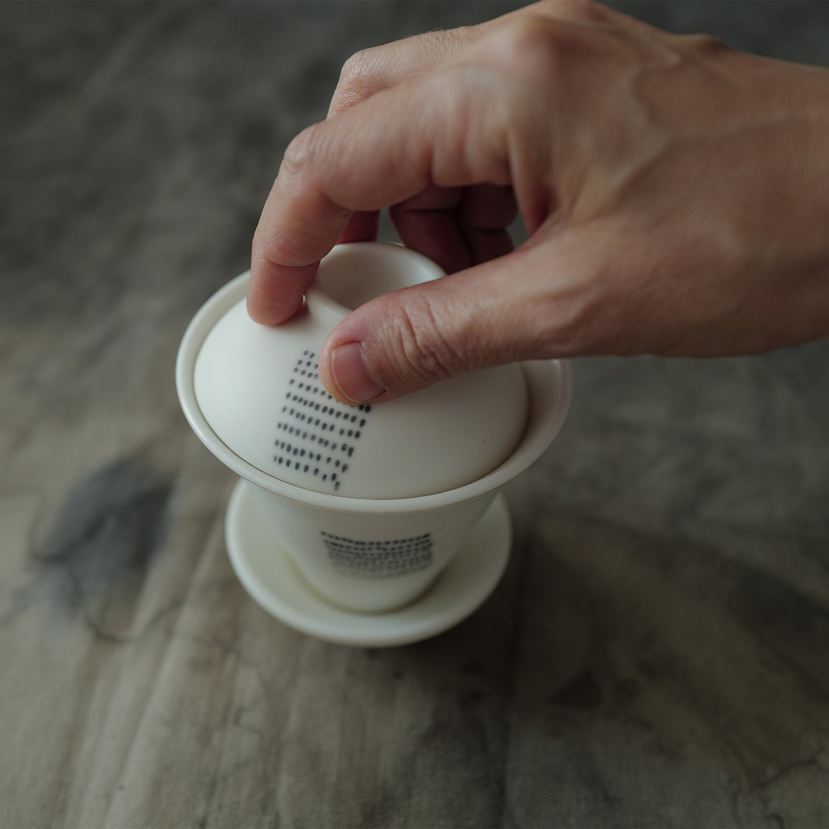 Hand holding a white ceramic gaiwan with a dotted pattern on a textured surface.
