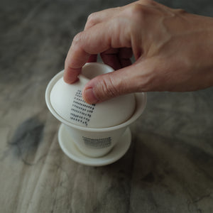 Hand holding a white ceramic gaiwan with a dotted pattern on a textured surface.