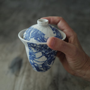 Hand holding a blue and white ceramic gaiwan with floral patterns against a blurred background.