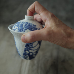 Hand holding a blue and white ceramic gaiwan with floral patterns against a blurred background.