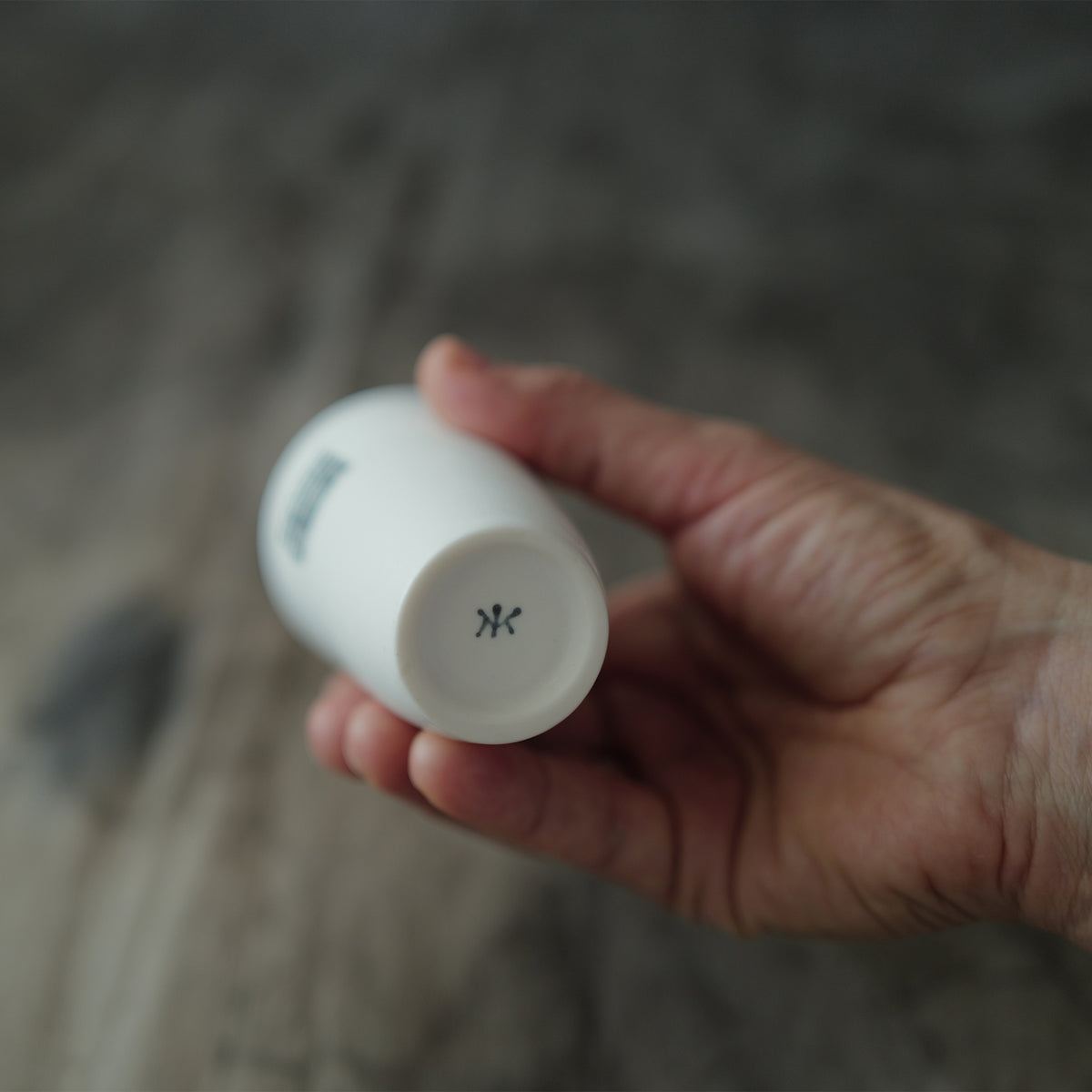 Hand holding a white ceramic teacup with a potter's mark against a blurred natural background.