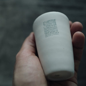 Hand holding one white  teacup with a textured surface against a blurred background.