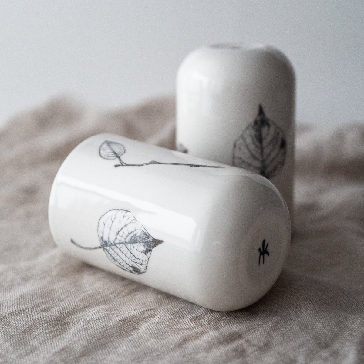 Two white porcelain tumblers with floral and twig designs, placed on a textured surface.